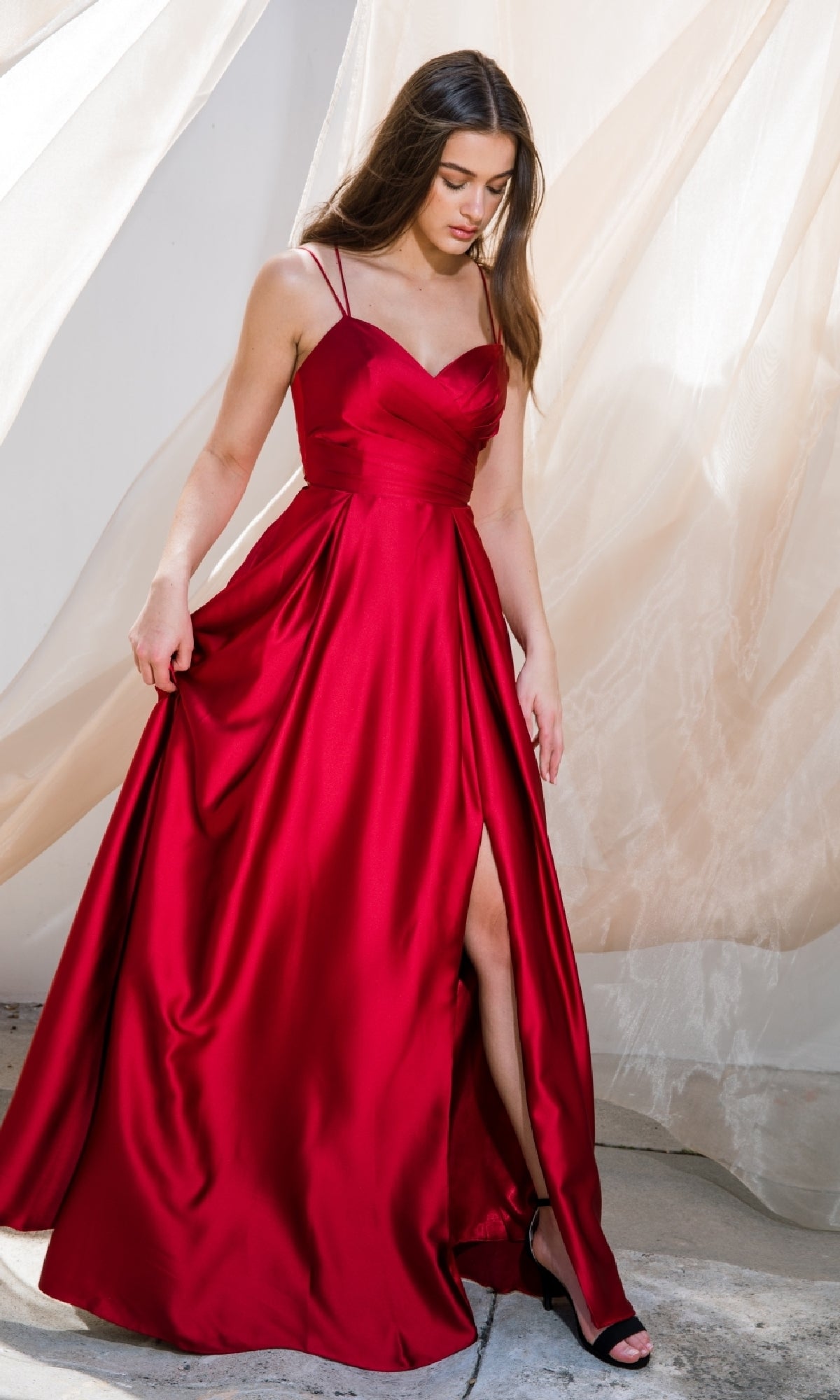 A person in the Strappy-Back Long Satin Prom Dress: Eureka 9925, featuring thin straps and a thigh-high slit, stands on a concrete floor near sheer curtains.
